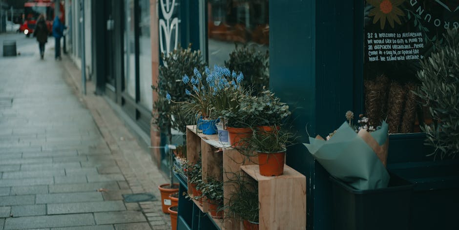 A row of potted flowering plants arranged on wooden shelves outside a flower shop located next to Enfield Town Station. The plants include a variety of colors, with some featuring blue, purple, and wh