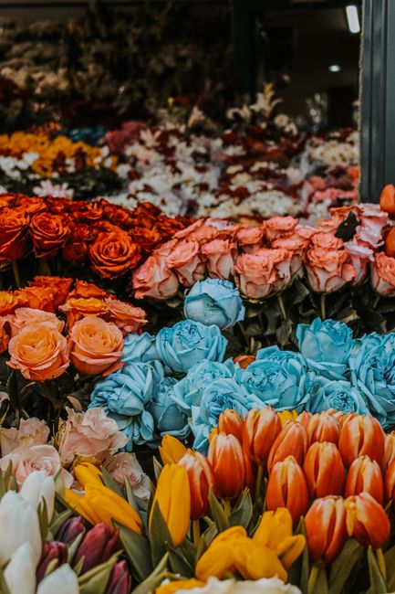 A vibrant display of fresh flower bouquets arranged in a floral shop. In the foreground, there are orange and yellow tulips with smooth, elongated petals. Behind them, clusters of blue roses with tigh